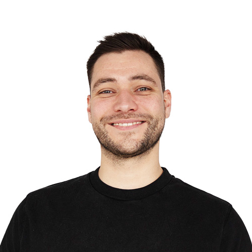 Ein lächelnder junger Mann mit dunkelbraunen, kurzen Haaren, einem Drei-Tage-Bart und einem schwarzen Pullover.