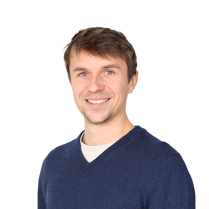 Ein lächelnder, junger Mann mit dunkelbraunen Haaren, blauen Augen, einem Drei-Tage-Bart, einem dunkelblauen Pullover und darunter einem weißen Oberteil.