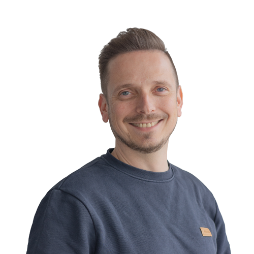 Ein lächelnder junger Mann mit hellbraunen, kurzen Haaren, einem Drei-Tage-Bart und einem dunkelblauen Oberteil.