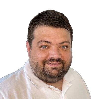 Ein Mann mit dunkelbraunen Haaren, einem dunkelbraunen Bart und einem weißen Oberteil.