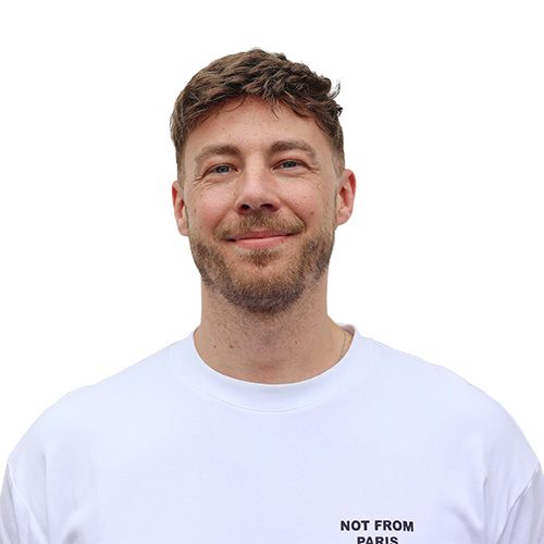 Ein lächelnder, junger Mann mit hellblonden Haaren, einem Drei-Tage-Bart und einem weißen T-Shirt.
