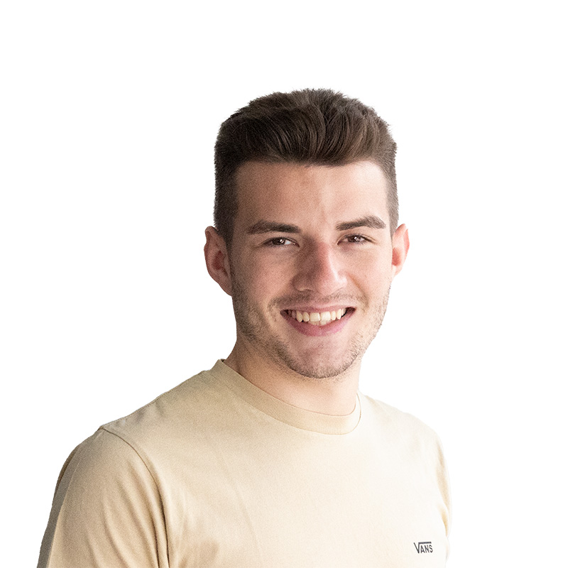 Ein lächelnder, junger Mann mit dunklen Haaren, einem Drei-Tage-Bart und einem beigen T-Shirt mit einem schwarzen Schriftzug.