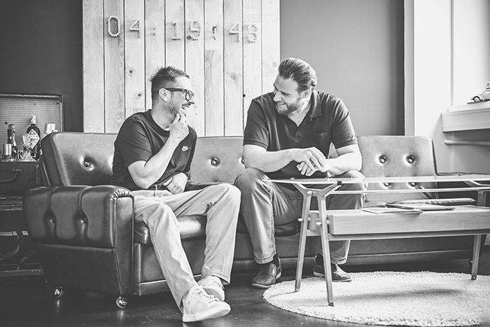 Zwei Personen sitzen in einem modernen Lounge‑Bereich auf Ledersofas und beugen sich über einen niedrigen Couchtisch, während sie sich miteinander unterhalten. Im Hintergrund steht eine Holzwand mit großen Zahlen, daneben verschiedene Dekorationselemente.